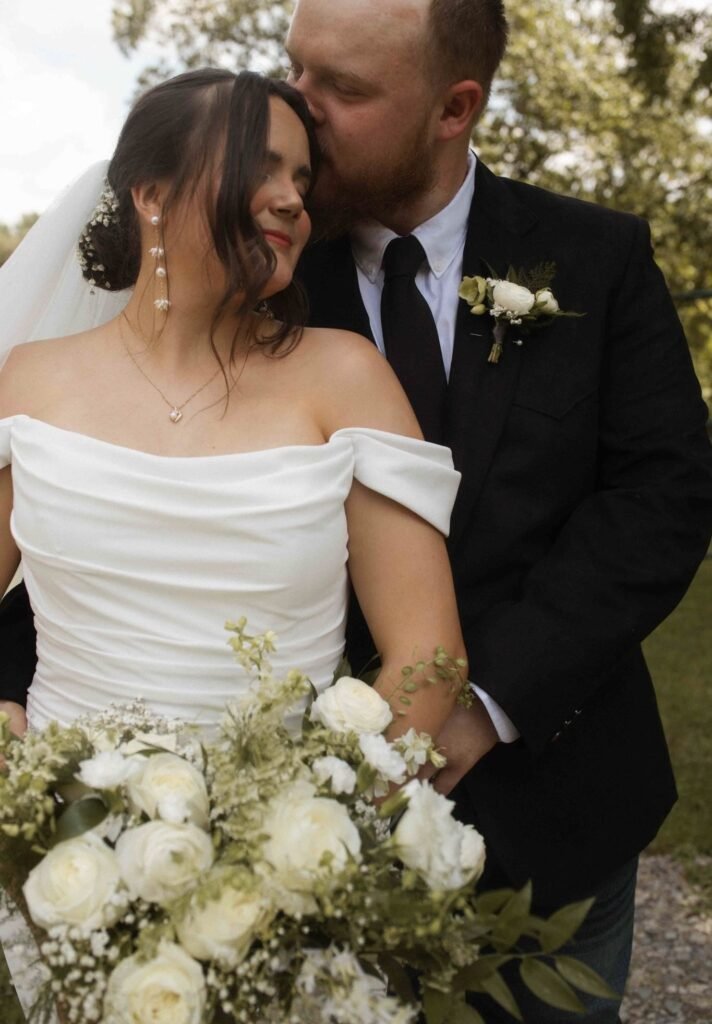 A bride holding a bouquet of flowers while a groom kisses her on the head.
