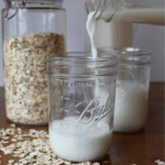 Oat milk being poured into a glass surrounded by oats on the table.