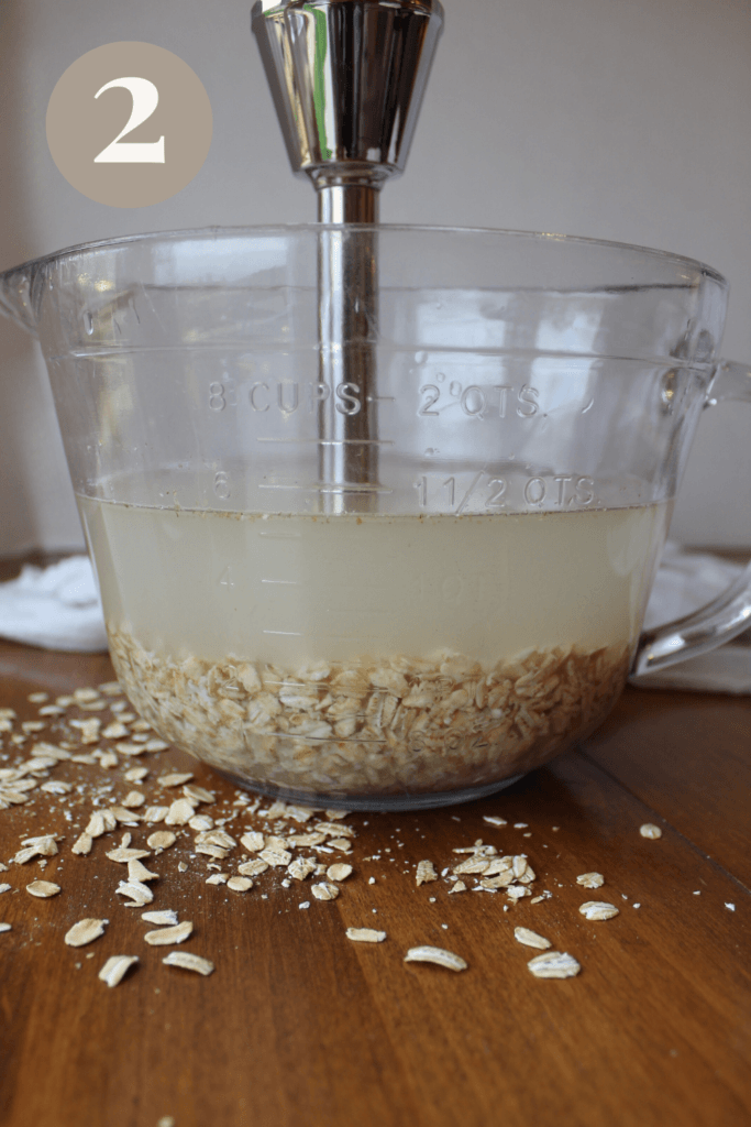 Oats and water in a glass bowl with an emersion blender in the bowl.
