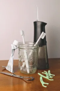 Natural oral care setup featuring a mason jar holding two white toothbrushes, a crumpled toothpaste tube, and a water flosser in the background. A stainless steel tongue scraper and several mint-green floss picks are arranged on a wooden surface.
