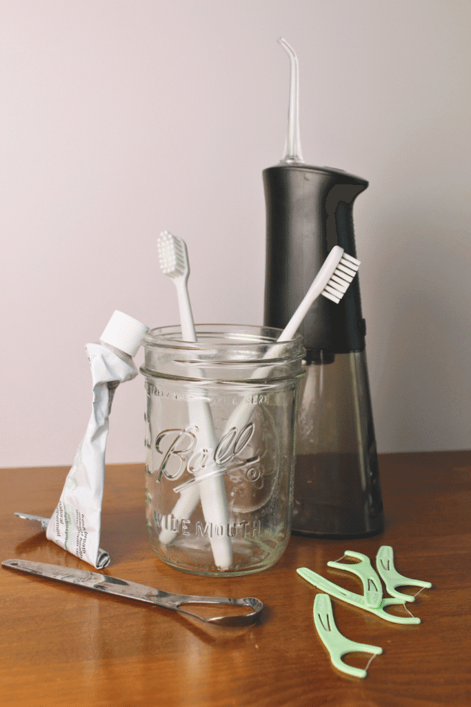 Natural oral care setup featuring a mason jar holding two white toothbrushes, a crumpled toothpaste tube, and a water flosser in the background. A stainless steel tongue scraper and several mint-green floss picks are arranged on a wooden surface.