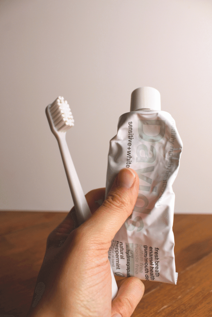 Close-up of a hand holding a white toothbrush and a crumpled tube of Davids natural nano-hydroxyapatite toothpaste with peppermint flavor, positioned against a simple neutral background.